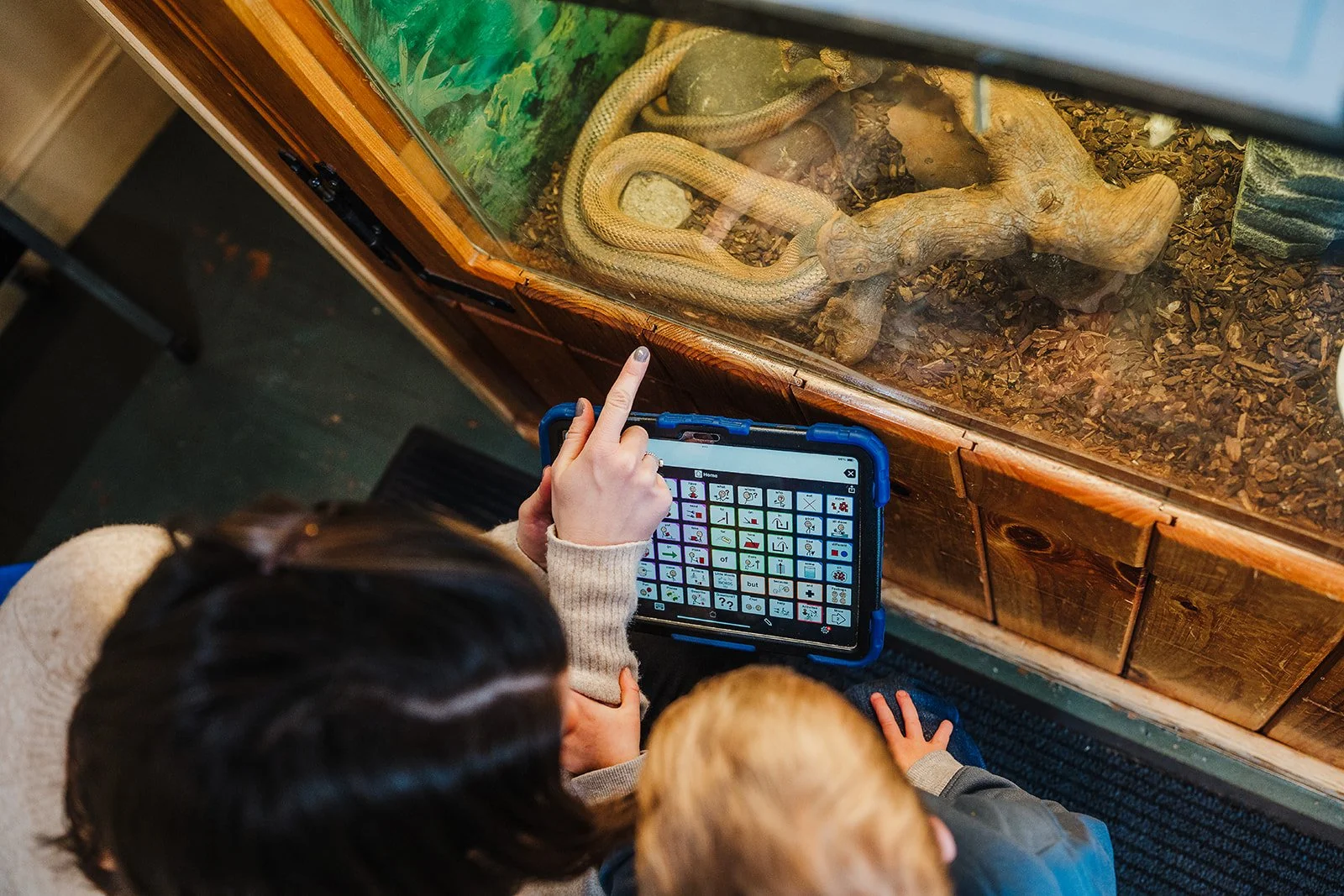 A woman and a young boy look at a snake while using a tablet AAC device to talk about it.  The boy is resting his hand on the woman's arm.