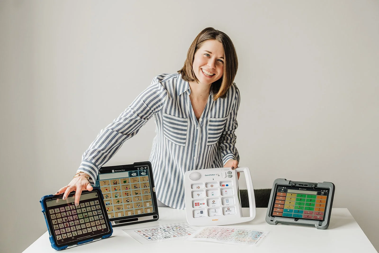 Woman with short brown hair is smiling while looking at the camera.  She is engaging with three AAC devices displayed on the table in front of her.