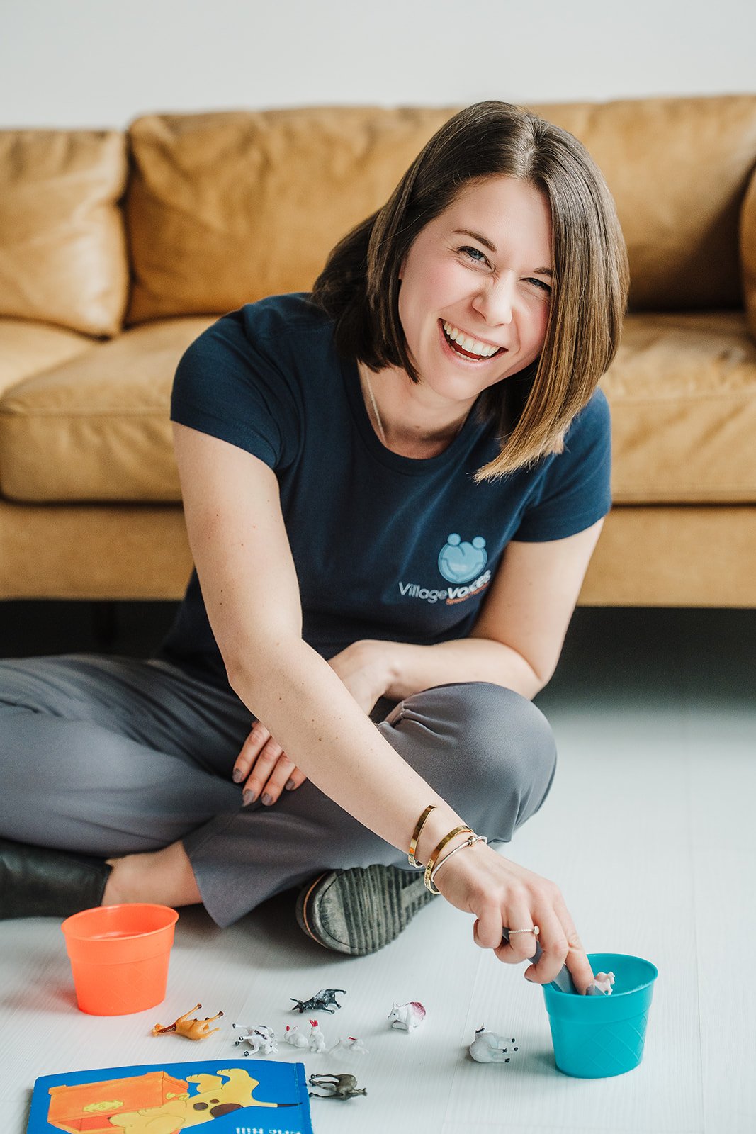 A woman with short brown hair is sitting cross-legged on the floor while playing with children's toys. She is smiling directly into the camera.
