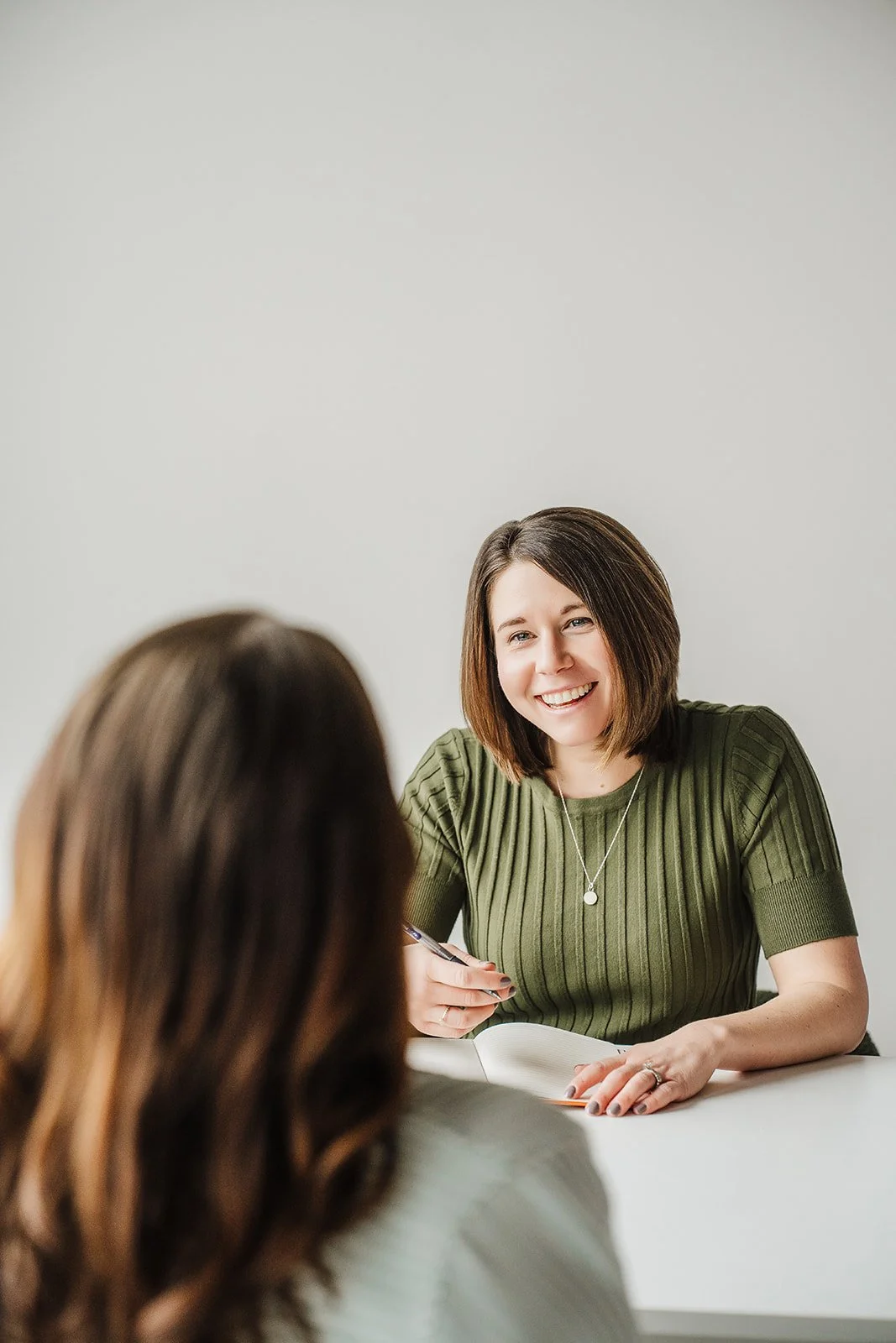 A woman with short brown hair is smiling and writing in a notebook while seated in front of a client.  The back of the clients head is shown as she faces Elena.