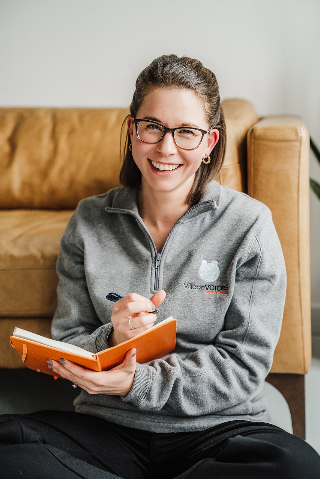 A woman is smiling at the camera while writing in an orange notebook.  She is wearing glasses and a gray Village Voices Speech Therapy sweatshirt.