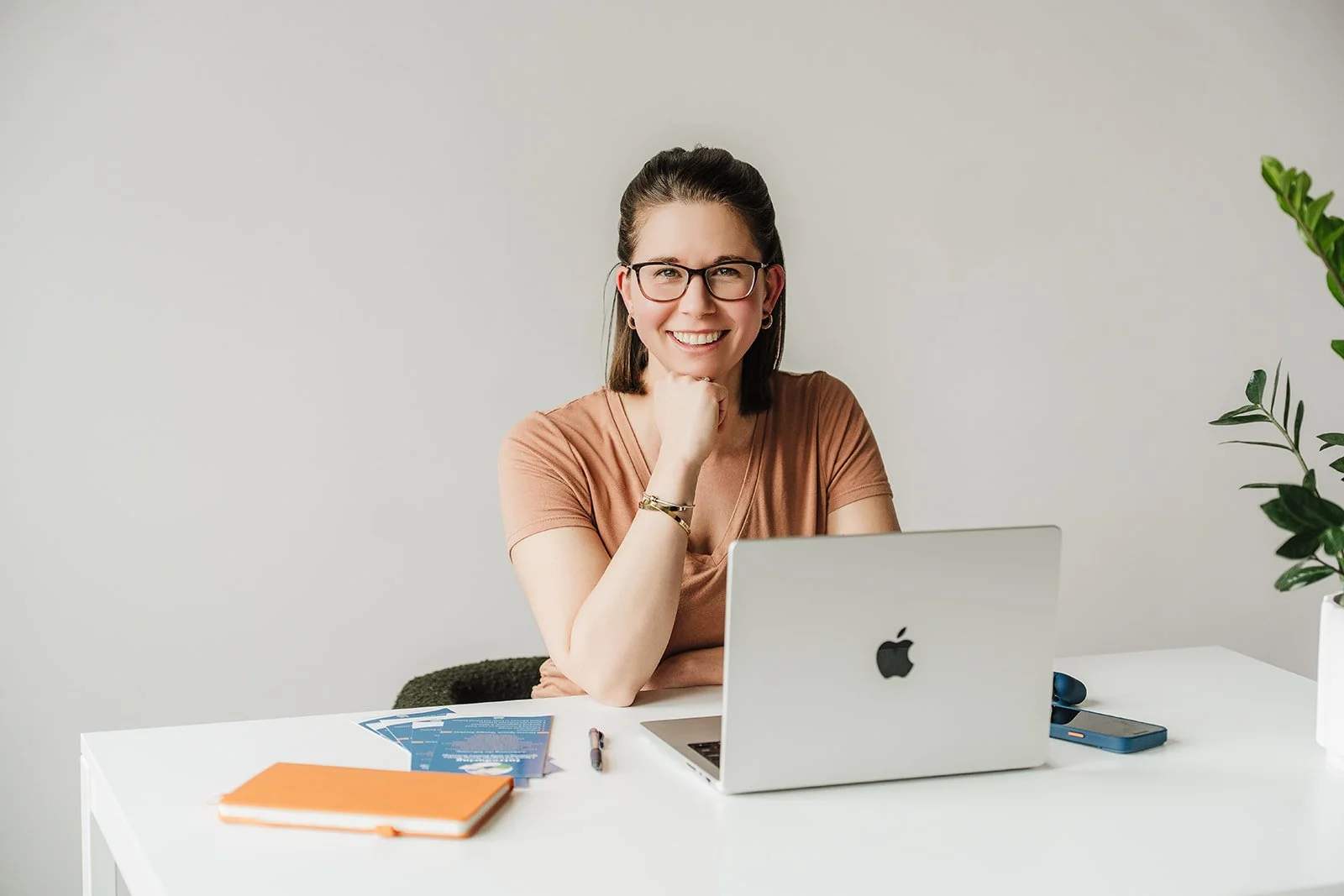 A woman with short brown hair and wearing glasses is seated in front of a MacBook while smiling and resting her head in her right hand.