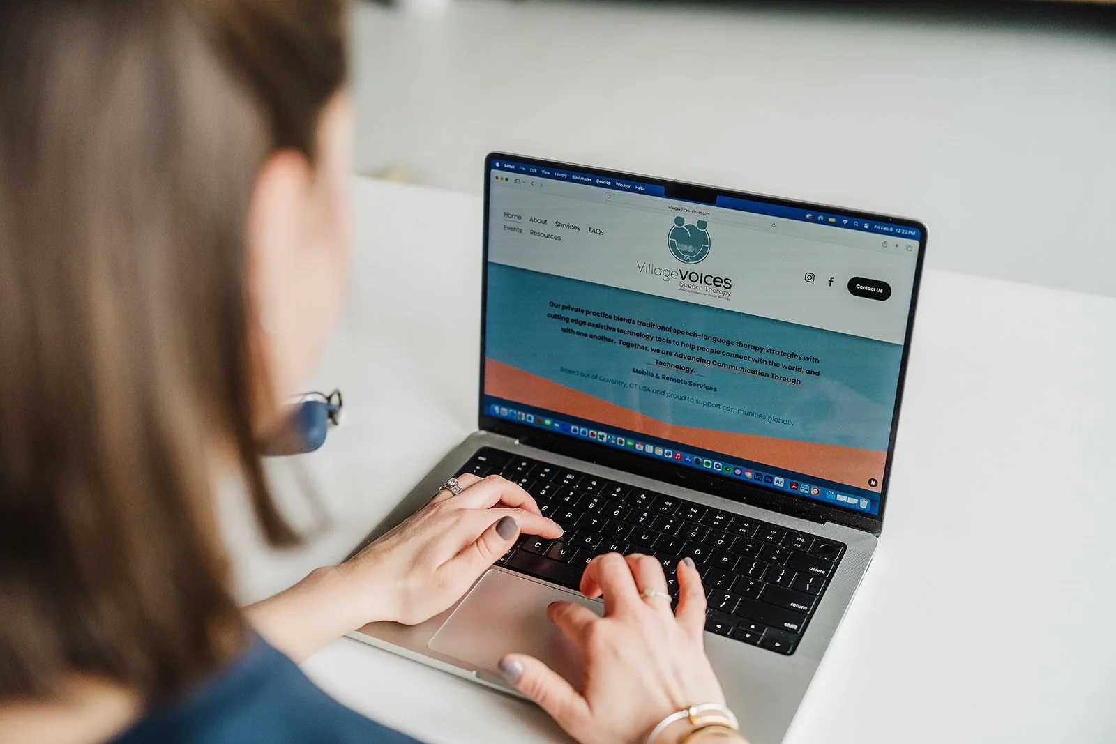The back of a woman's head is blurred with the image focused on her typing on a laptop with the Village Voices Speech Therapy website displayed