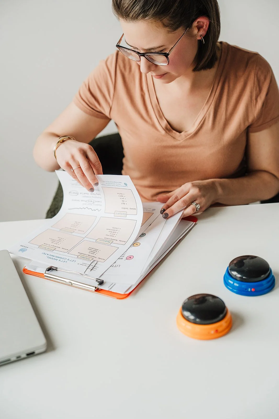 A woman looking down at a clipboard and shuffling through paper resources.  Two mid-tech AAC buttons are next to her.