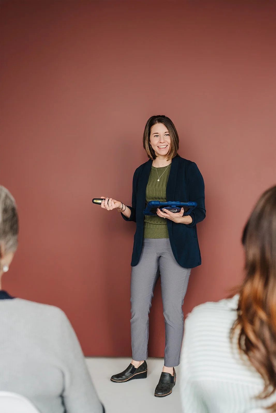 A woman giving a presentation in front of a pink wall, holding a remote control and a tablet, wearing a dark green top, navy blazer, gray pants, and black wedge shoes, with two audience members visible.