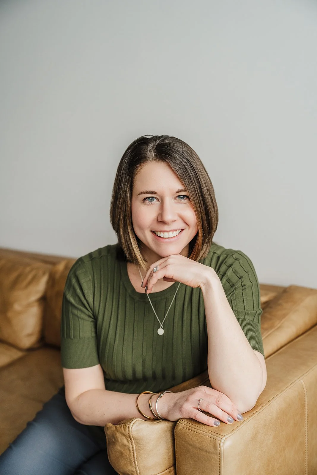 A woman with short brown hair wearing a green shirt and smiling at the camera while sitting on a tan couch.