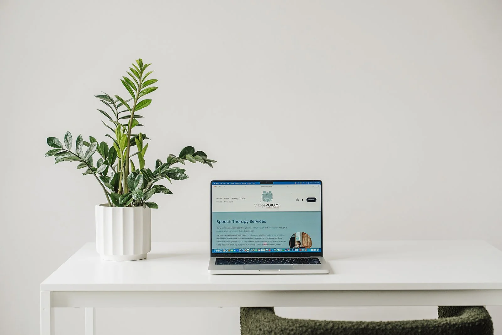 A laptop with the Village Voices Speech Therapy website displayed.  The laptop is on a white table and placed next to a green plant.