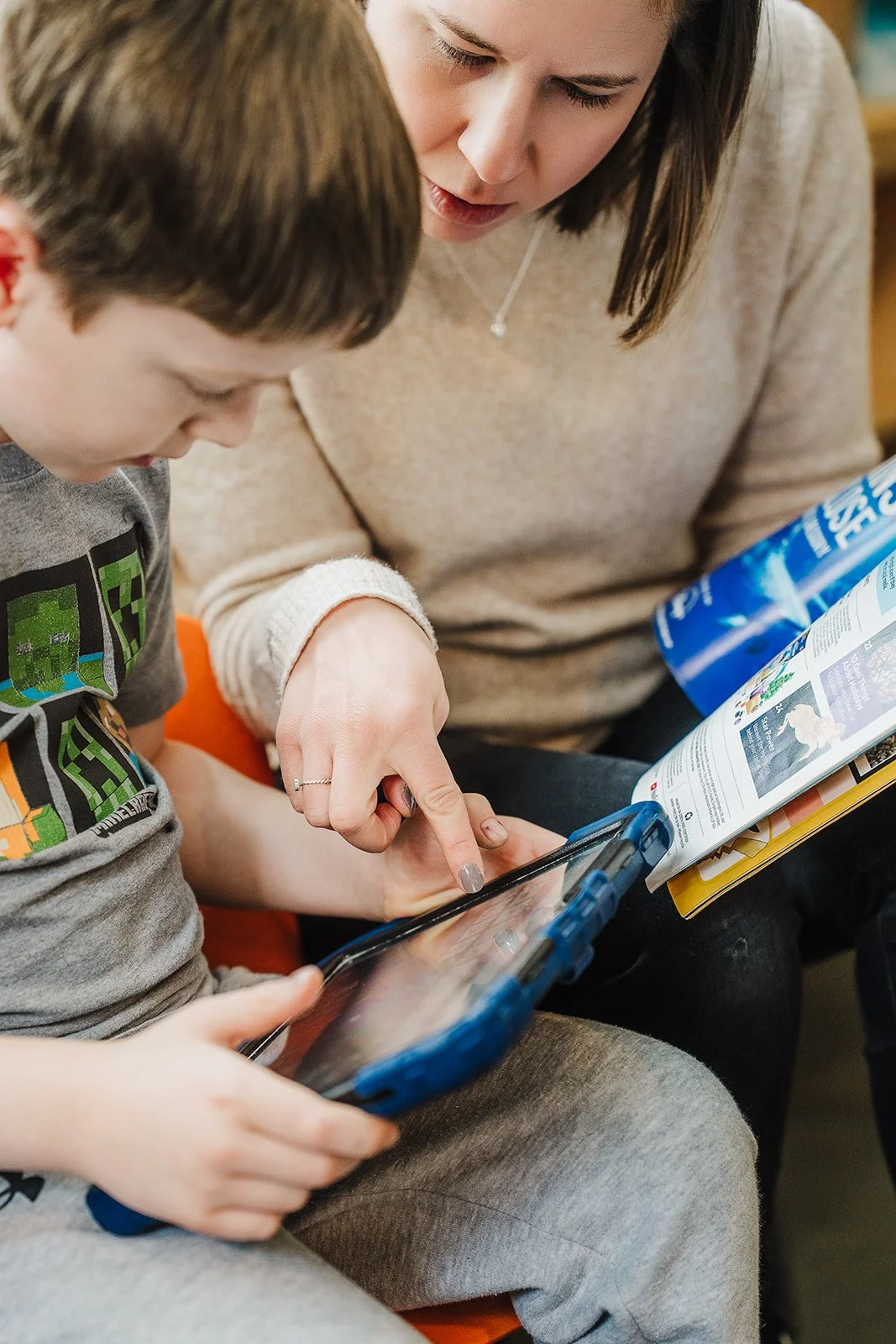 A woman and a young boy look at a magazine and an AAC device together
