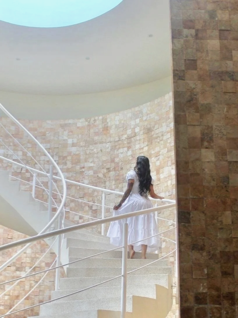 A woman with long black hair in a white dress walking up a curved staircase with a white railing, surrounded by a brick wall and a white ceiling with a circular cutout.