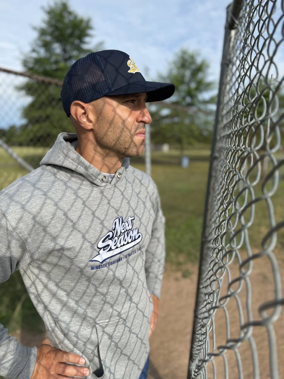 Man in gray hoodie and blue baseball hat standing on baseball field at fence. baseball mindset coach
