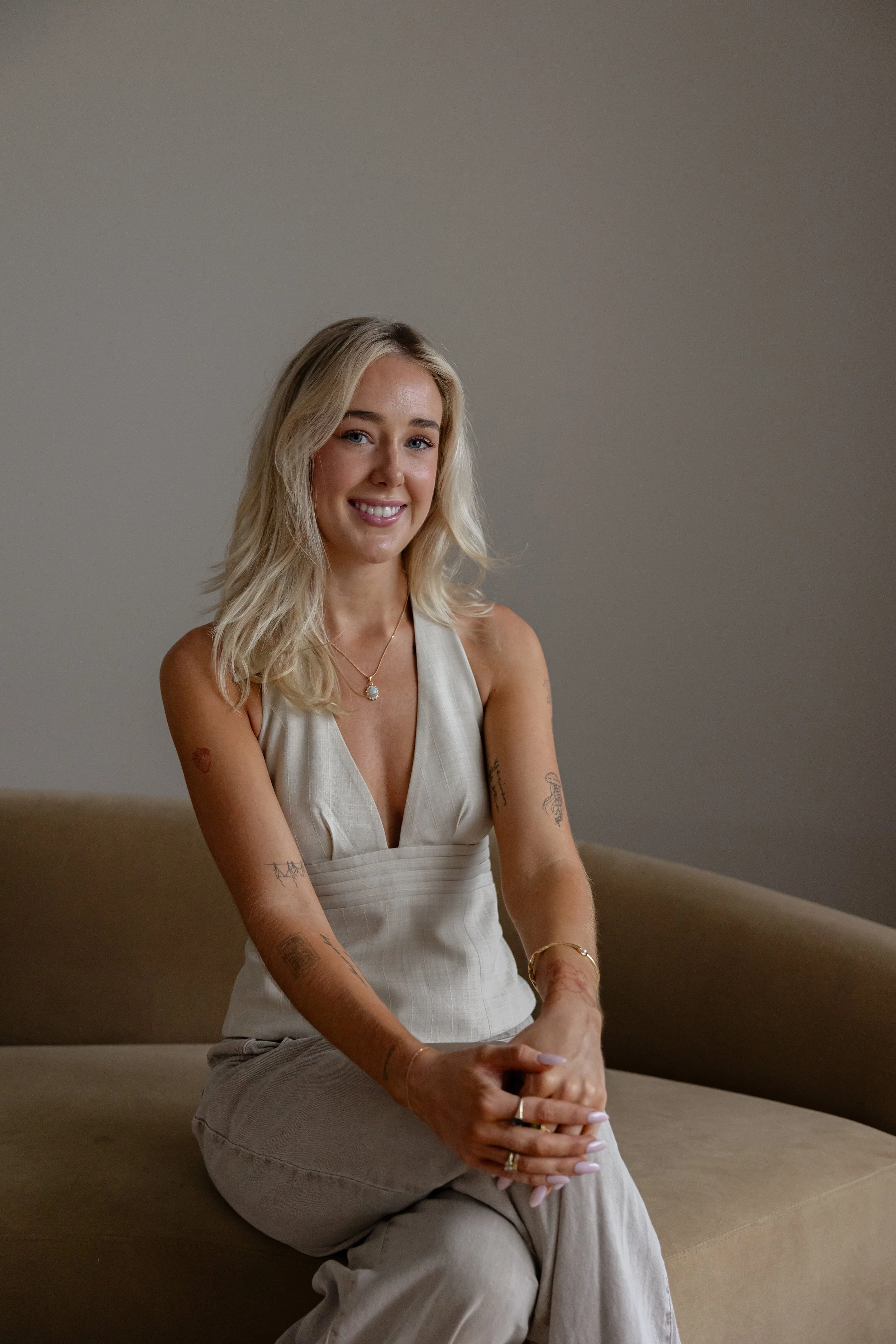 A young woman with blonde hair, blue eyes, and tattoos on her arms, sitting on a beige couch against a plain, light-colored wall. She is smiling, wearing a sleeveless beige top and matching pants, accessorized with a necklace, bracelets, and rings.
