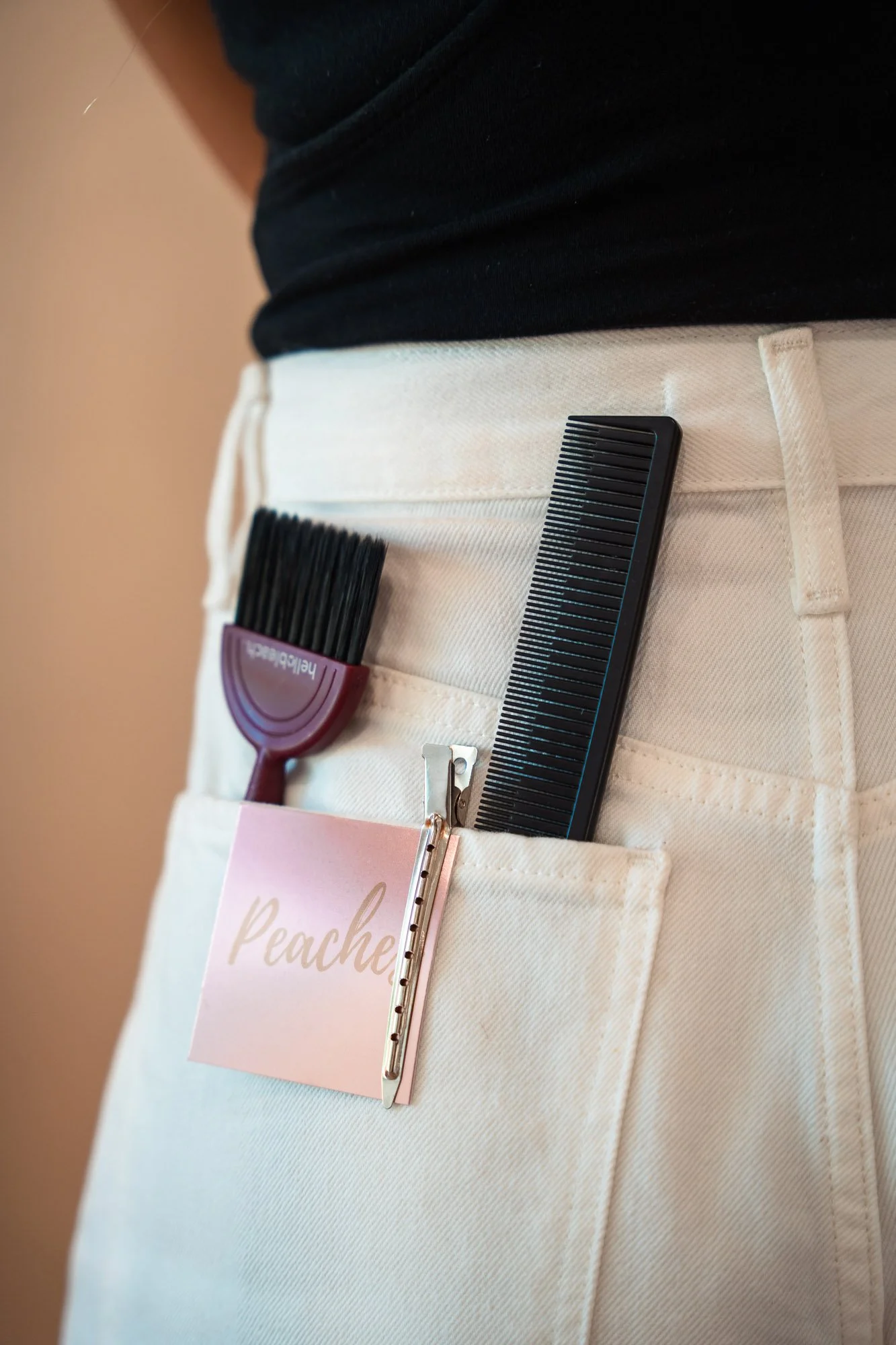 Close-up of a person's waist wearing beige pants, with a black comb, a pink nail file, a clothespin holding a pink tag labeled 'Peach', and a hairbrush in the front pocket.