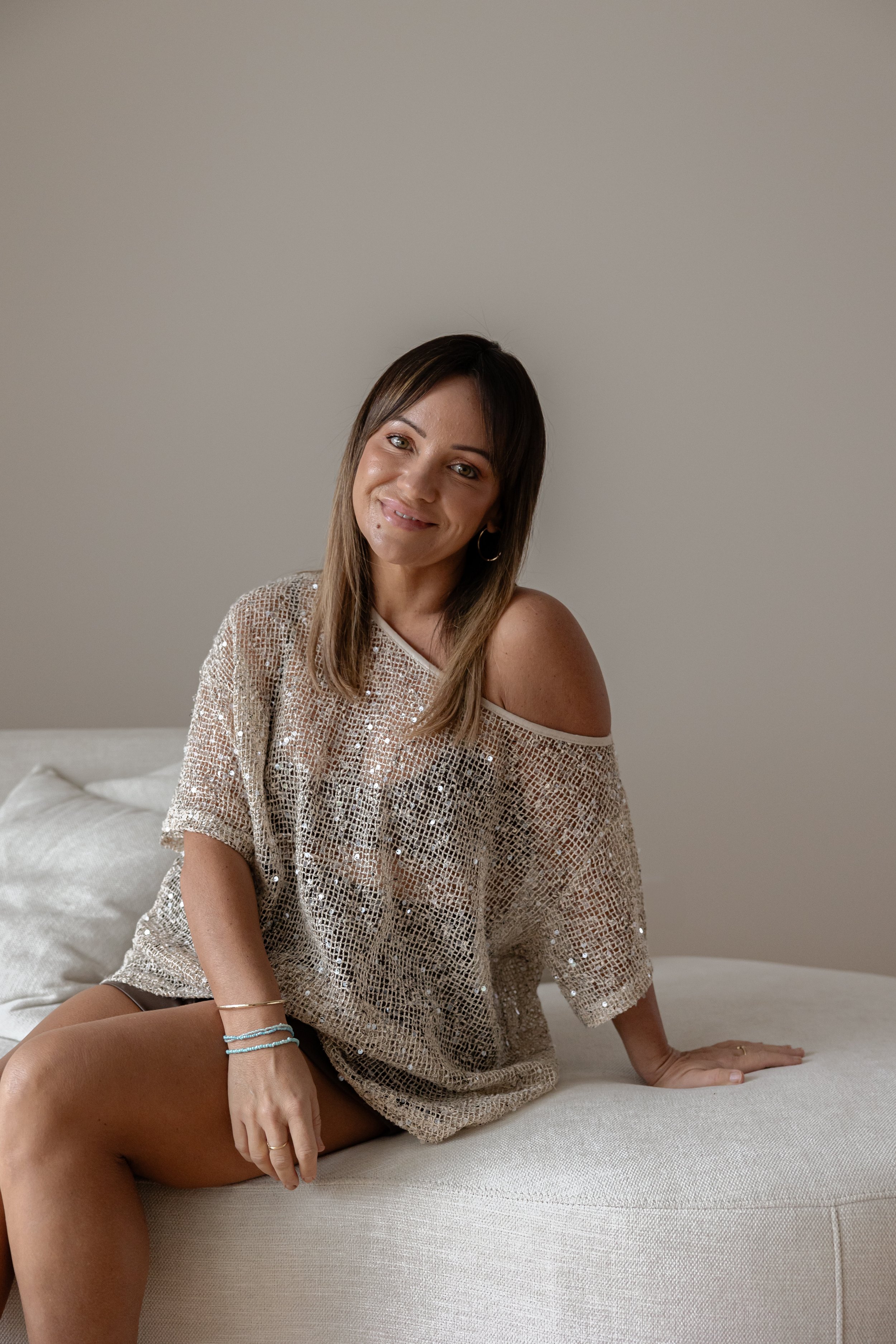 A woman with straight shoulder-length brown hair sitting on a white couch against a plain light-colored wall, wearing a loose, off-the-shoulder, semi-sheer, sparkly beige top, with multiple bracelets on her left wrist, smiling softly.