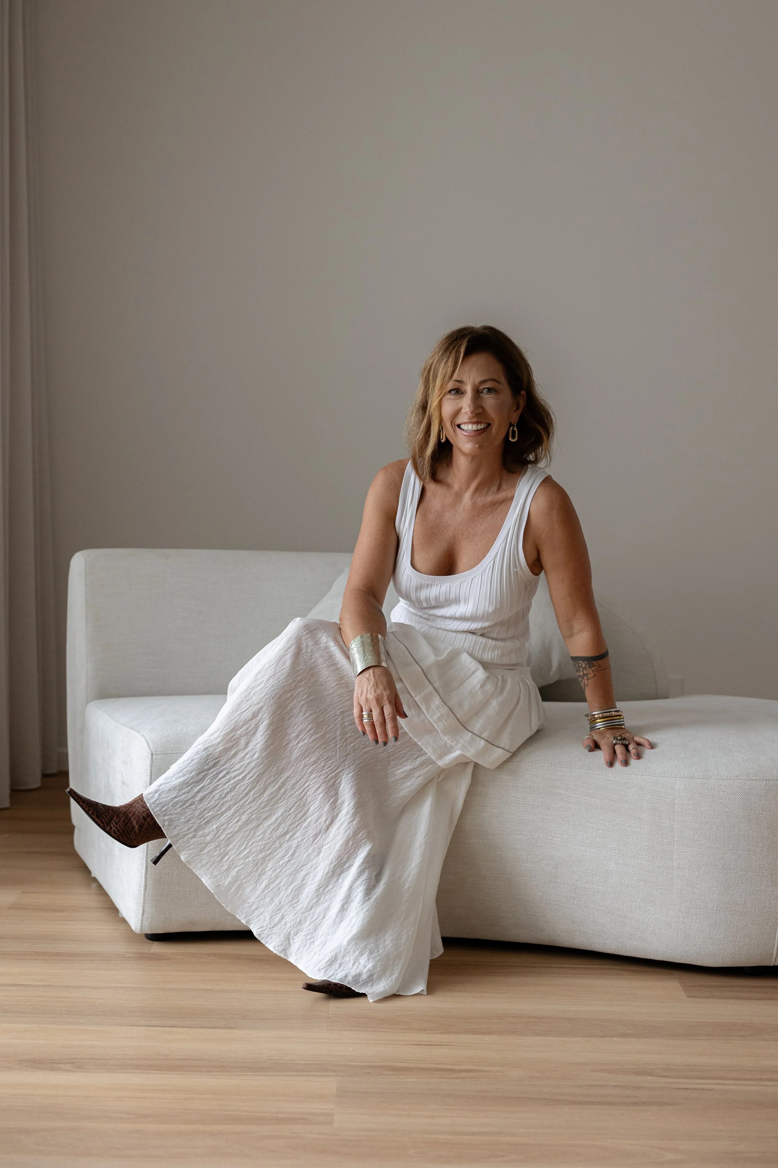 A woman sitting on a white sofa in a minimalist room, smiling, wearing a white sleeveless top and a long white skirt, with jewelry and bracelets, and brown pointed shoes.