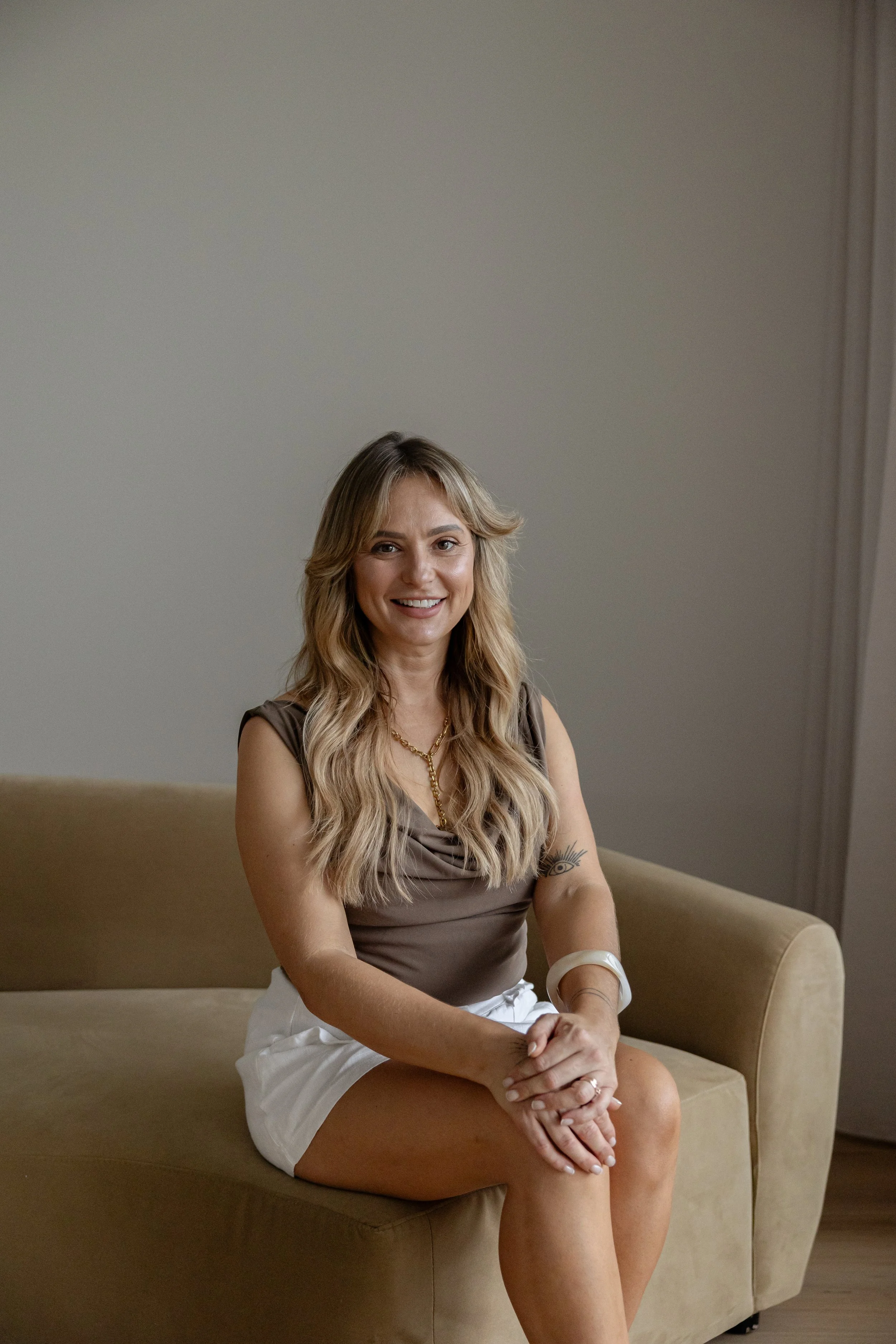 A smiling woman with wavy blonde hair, wearing a sleeveless brown top, white shorts, and gold jewelry, seated on a beige sofa in front of a plain wall.