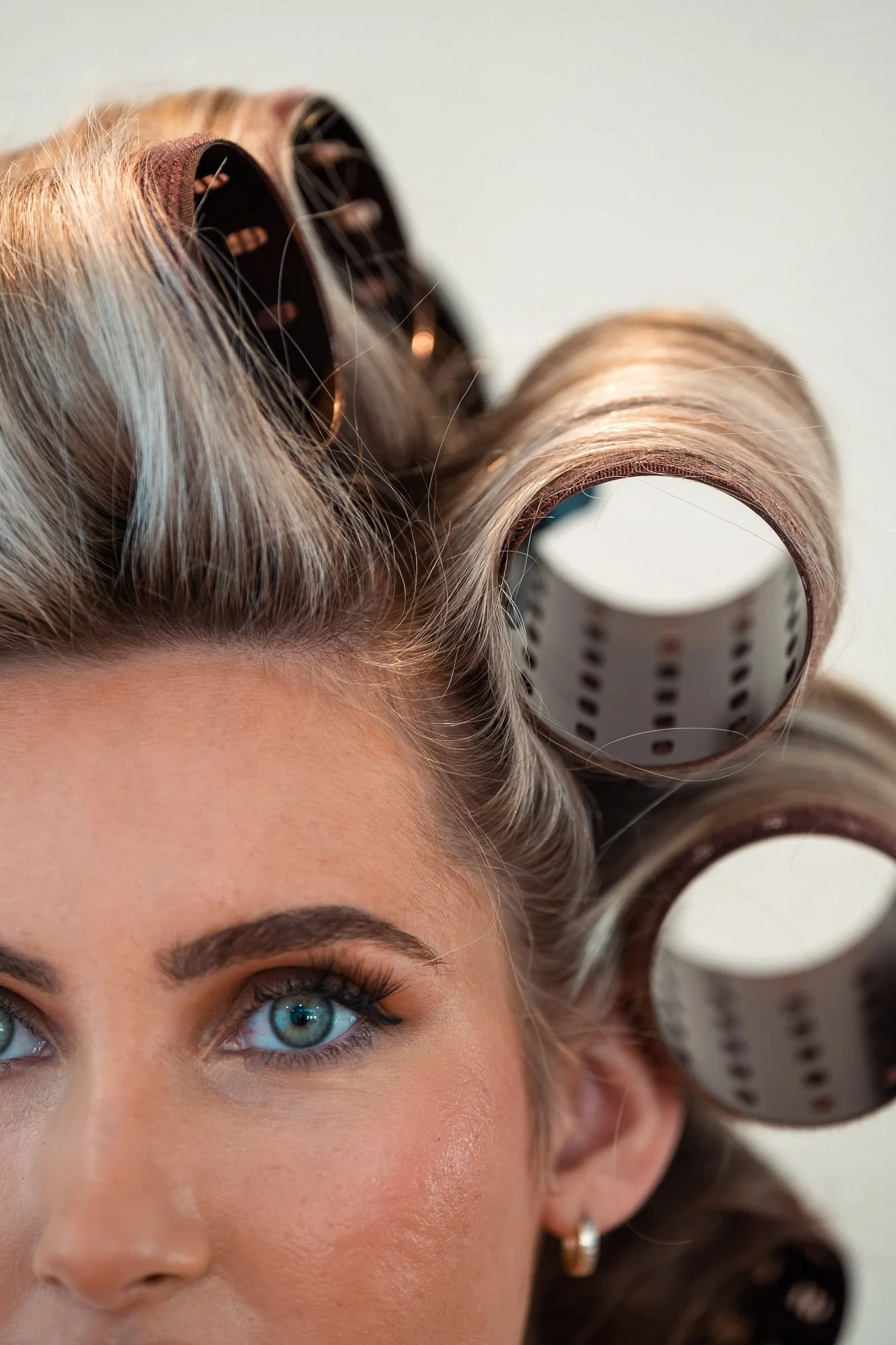 A woman with blue eyes and light makeup, hair in rollers, and an earring, looking upward.