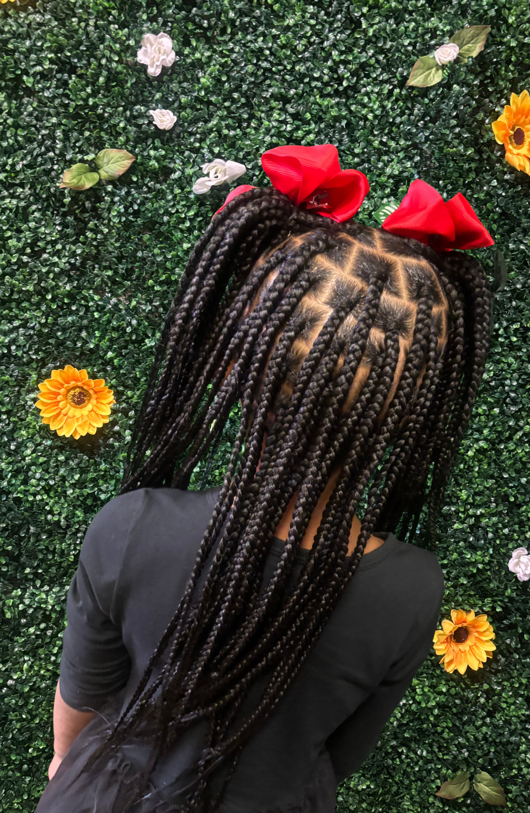 Back of person's head featuring long black box braids adorned with large red bows, standing in front of a green leafy wall with scattered artificial flowers.