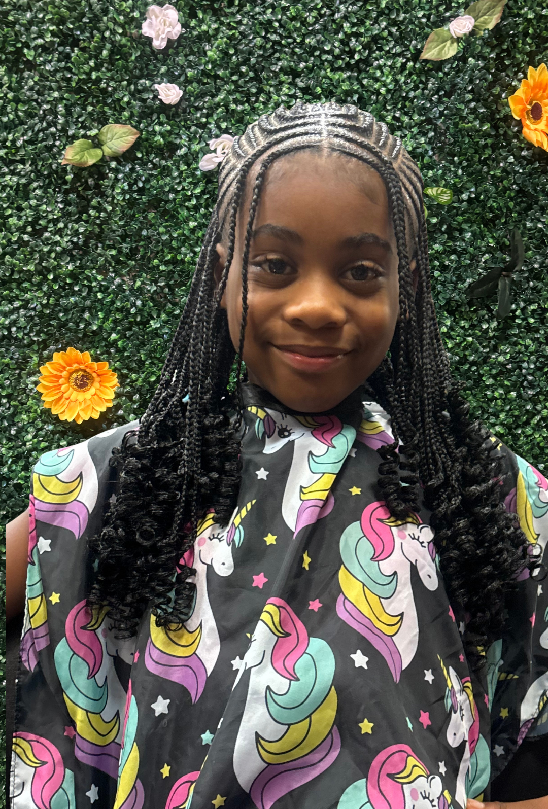 A young girl with braided hair standing in front of a green leafy wall decorated with artificial flowers and sunflowers wearing a cape with colorful unicorn and pastel rainbow patterns.