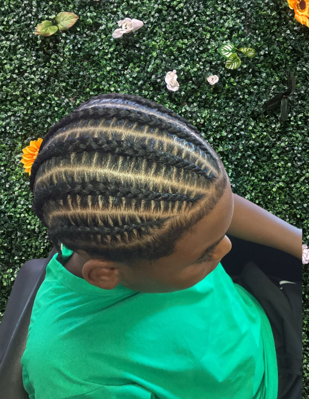 Top view of a person with cornrow braids decorated with blonde highlights, sitting on a black chair against a green leafy background with flowers.