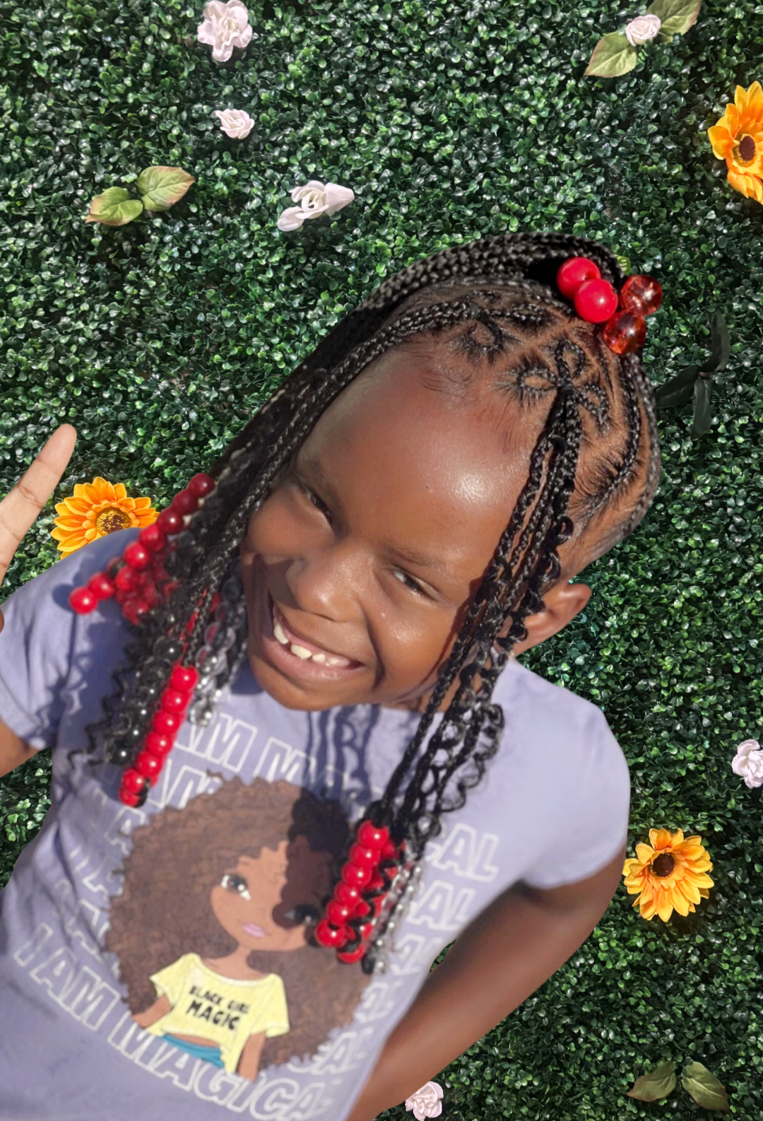 A smiling young girl with braided hair decorated with red, brown, and white beads, standing in front of a green leafy background with flowers.