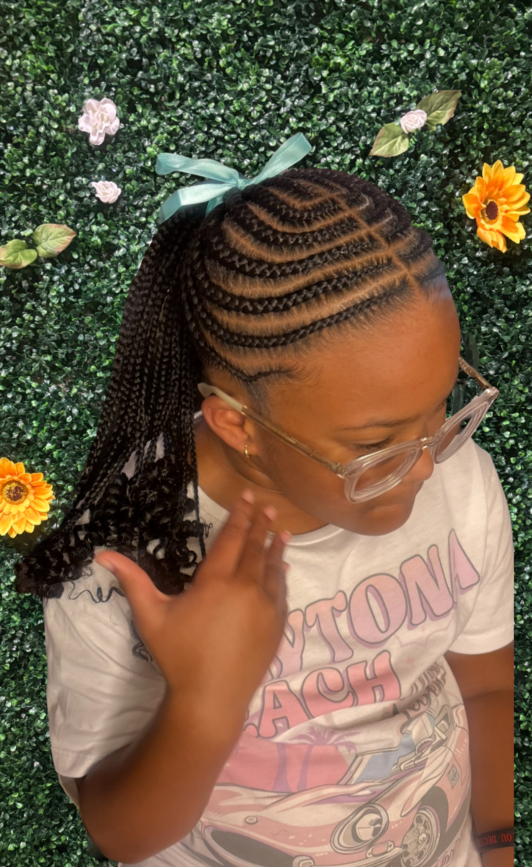 A woman with intricately braided hair, tied up with a light blue ribbon, touching her neck while wearing clear glasses and a T-shirt with a vintage car illustration, standing in front of a green leafy wall decorated with small flowers and a sunflower