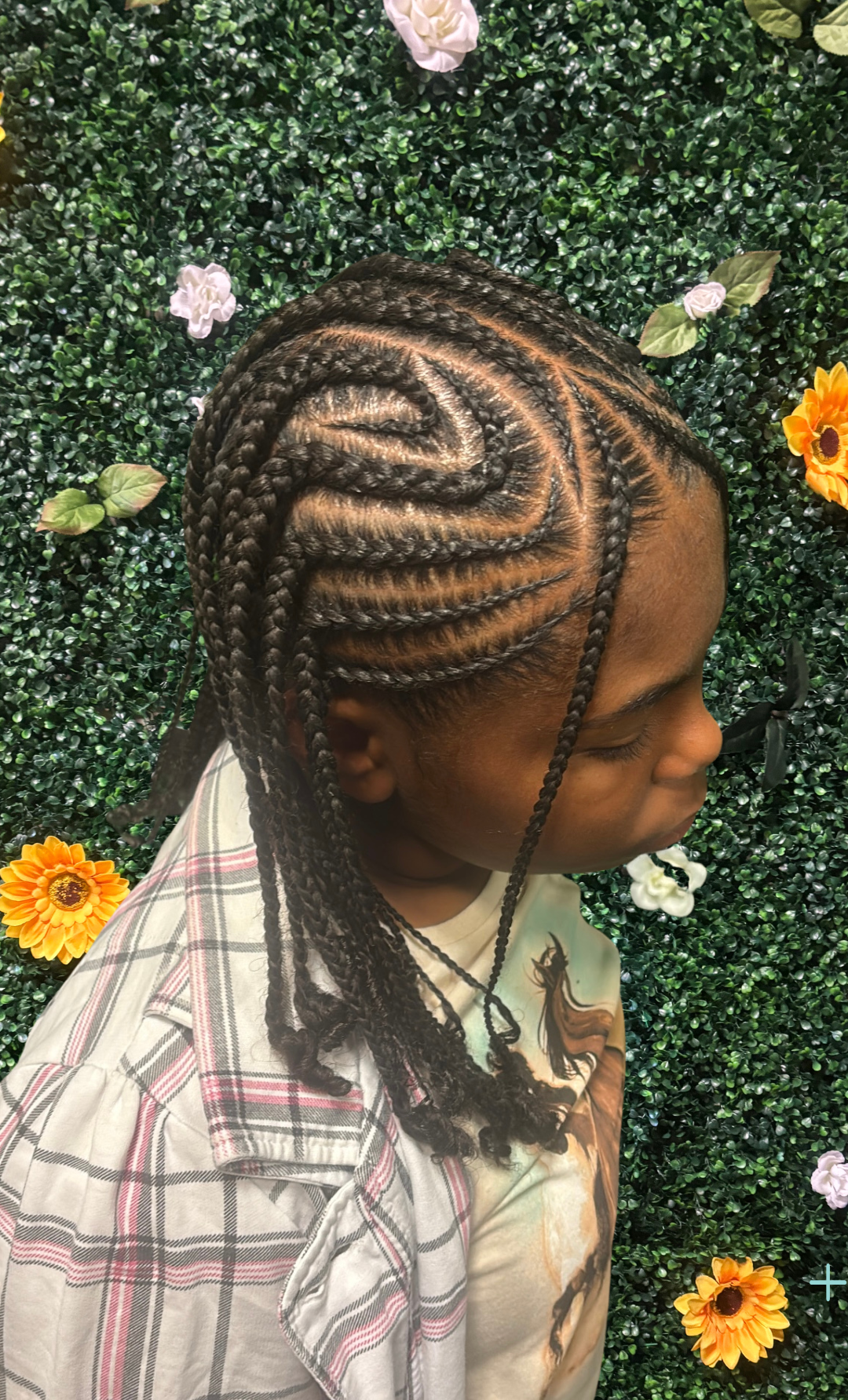 Child with braided hairstyle sitting in front of a green wall decorated with artificial flowers.