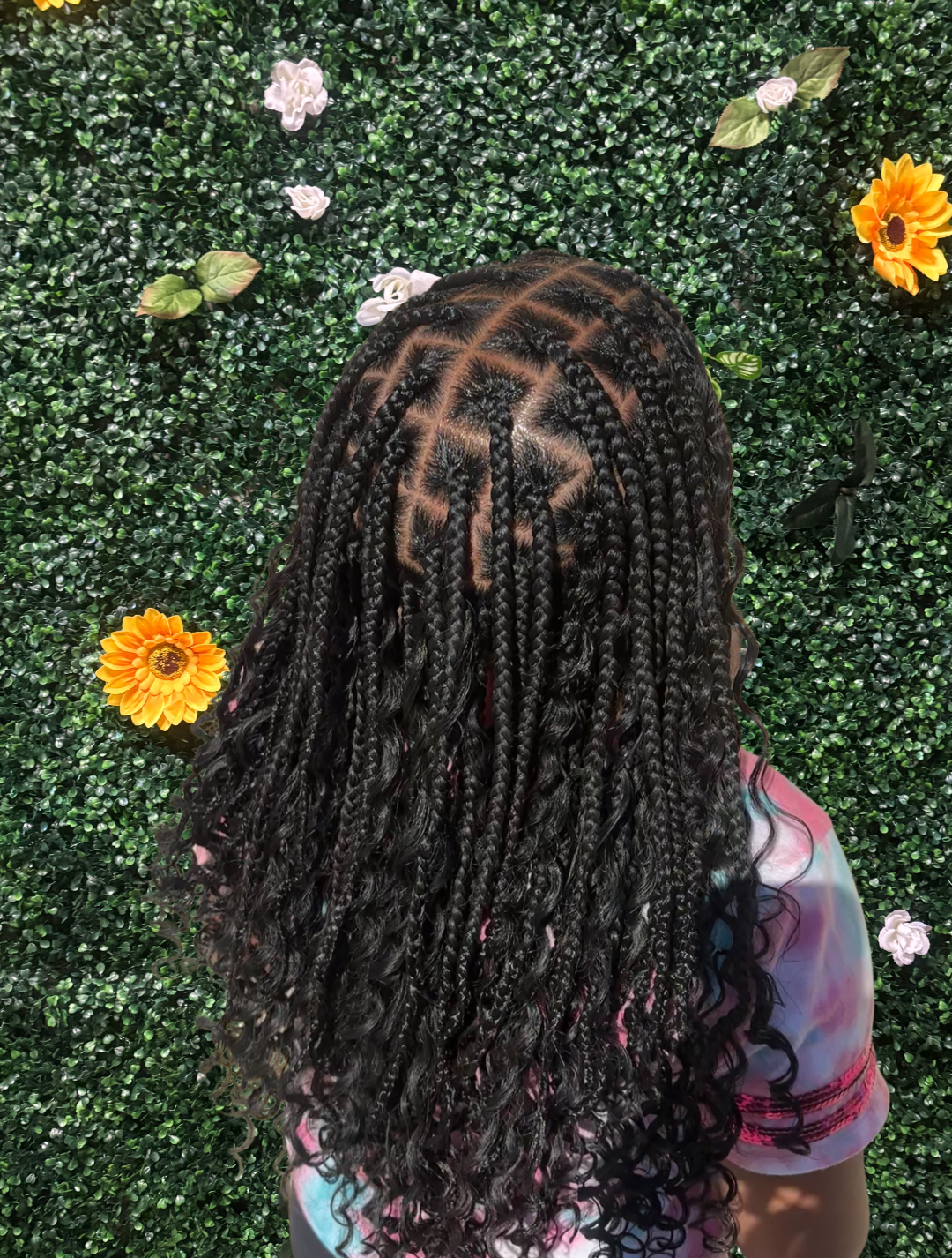 Back view of a person with tightly braided and curly black hair against a green leafy wall with flowers.