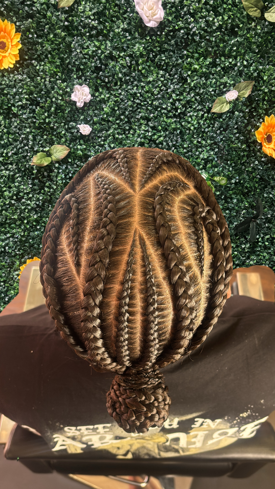 Top view of a person's braided hairstyle with cornrows and a braided bun at the nape, over a green leafy wall with flowers.
