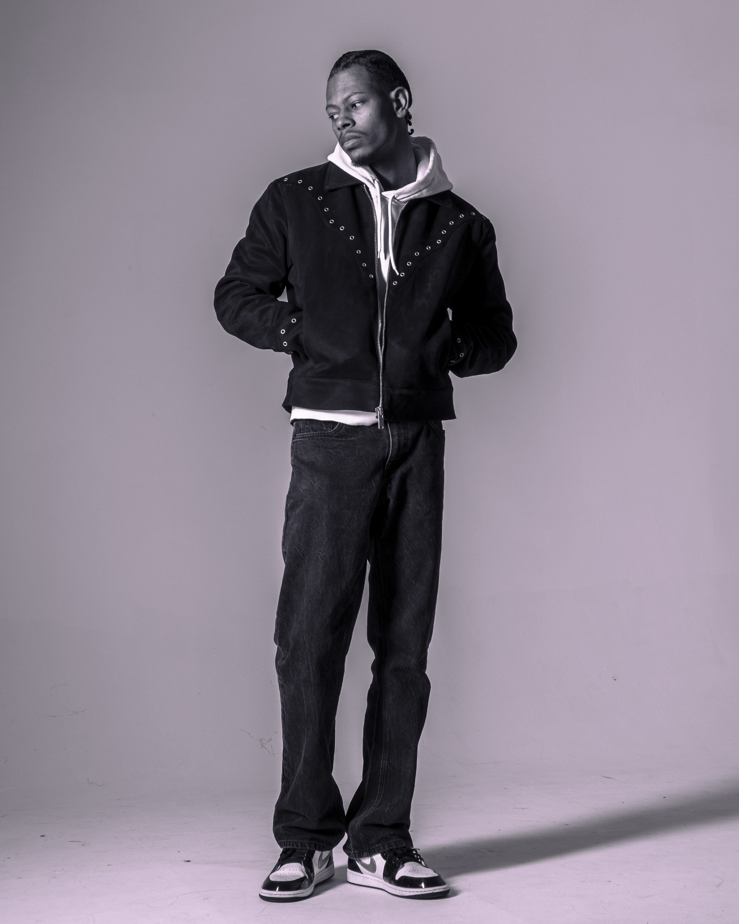 A man standing against a plain backdrop, looking slightly to his right with his hands in his pockets, wearing a black jacket with metal embellishments, a white hoodie, loose-fitting black jeans, and black and white sneakers.