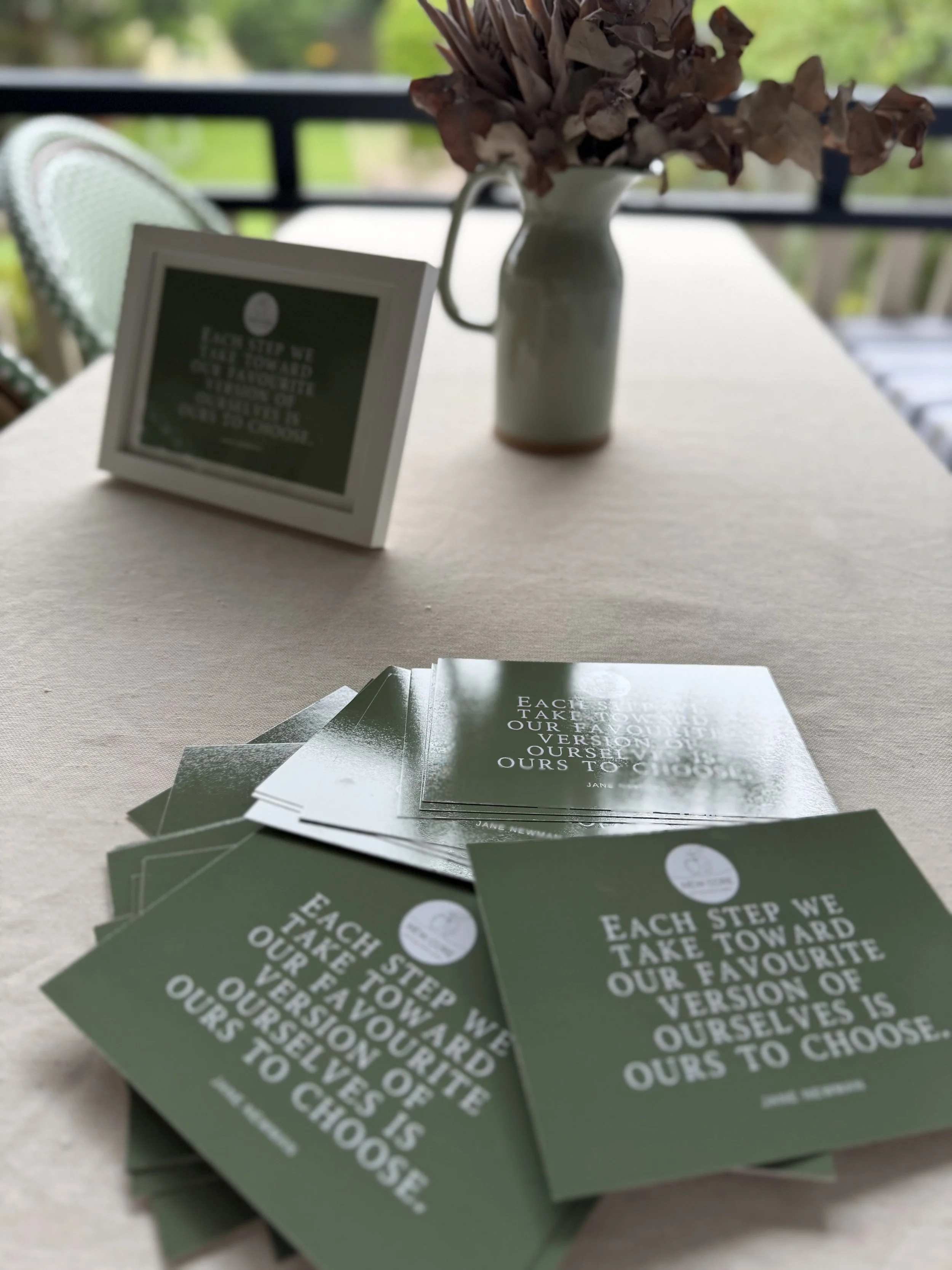 A table with a stack of dark green flyers or cards containing white text, a white vase with dried flowers, and a small blackboard or sign with additional text, set on a light-colored table outdoors, with a blurred background of greenery and balcony railing.