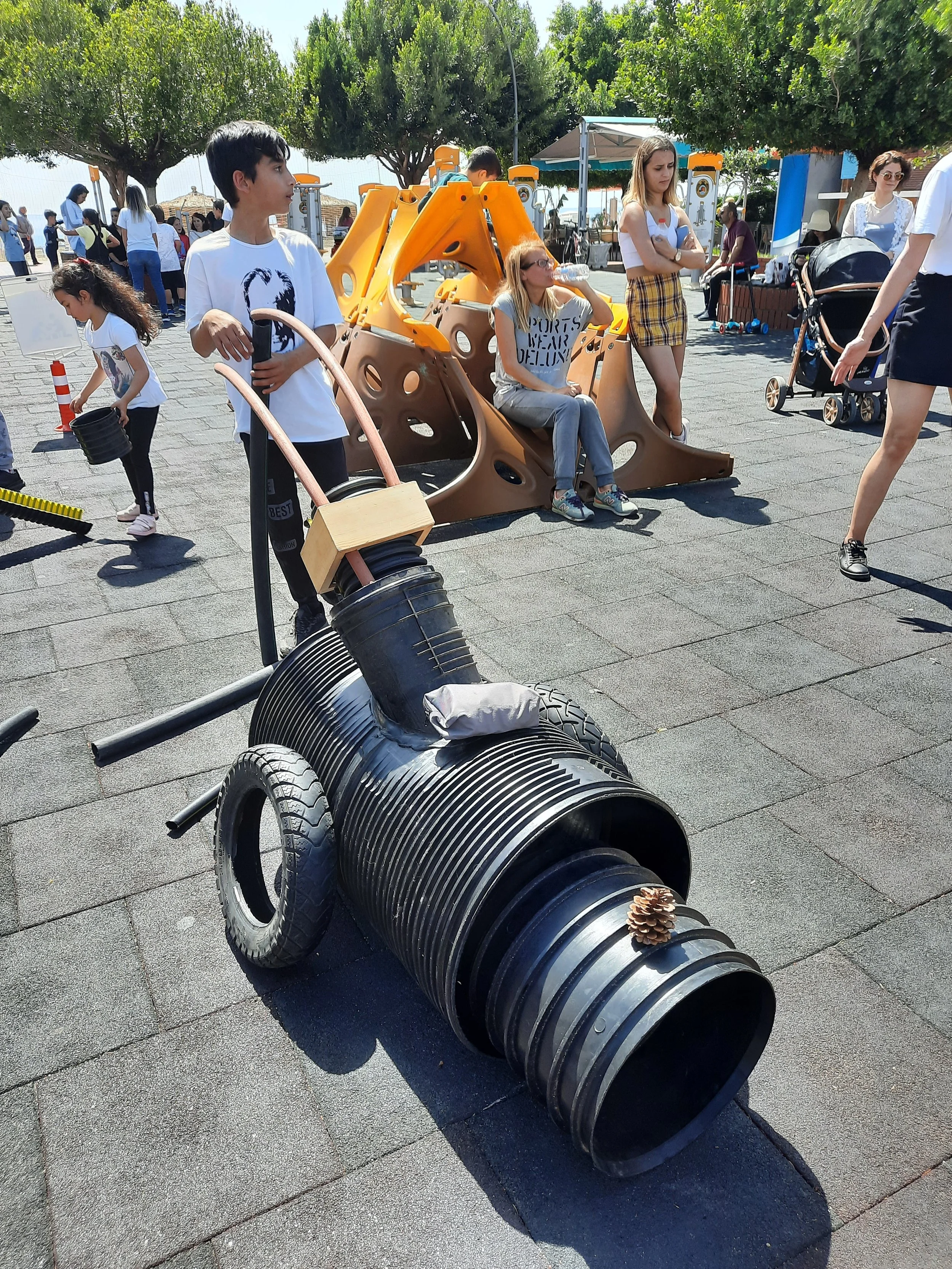 Children playing outdoors with recycled materials, including a robot made from large metal cans, and a playground structure in the background.