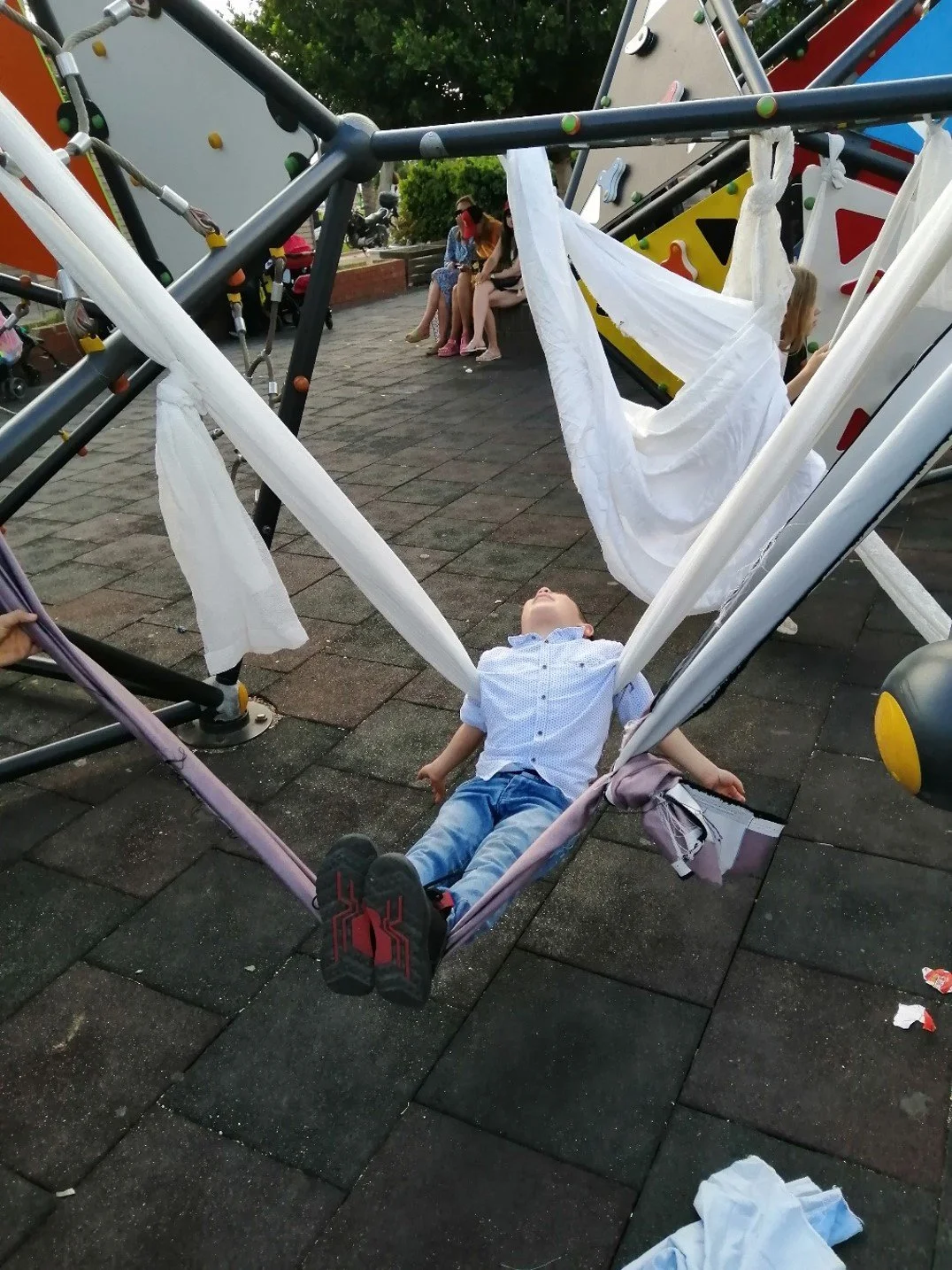 A young boy lying on a playground swing with fabric draped around him, looking up, with park benches and other children in the background.