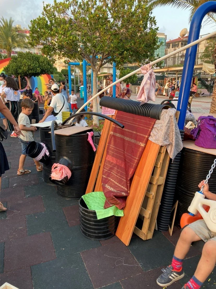 Children and adults at an outdoor market or festival with colorful stalls, trees, and buildings in the background. There is a makeshift structure with barrels and fabric in the foreground.