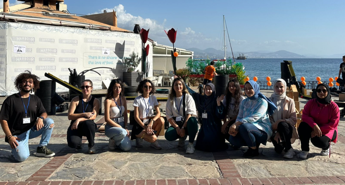 Ten people posing outdoors in front of a body of water with boats, some wearing name tags, with colorful decorations and a sign that reads "There is no scarier than the love of food."