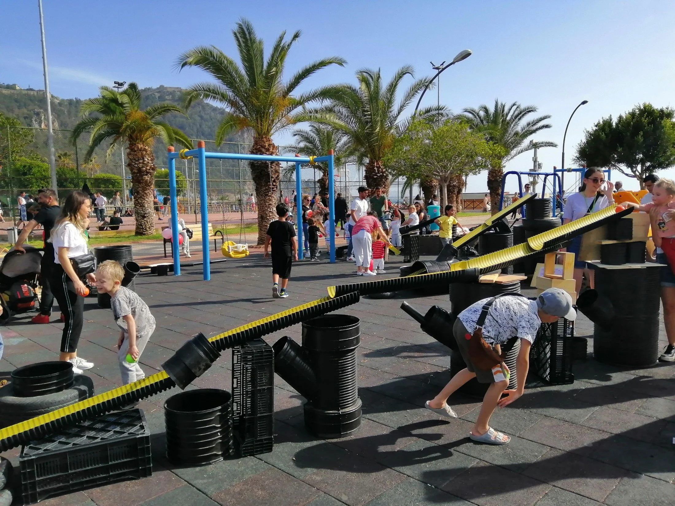 Children and adults in a park playing with a handmade Rube Goldberg machine on a paved area with palm trees and a playground in the background.