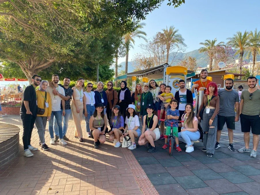 Group of people outdoors, some young children, standing and sitting around a small train ride at a park, with trees, mountains, and palm trees in the background.