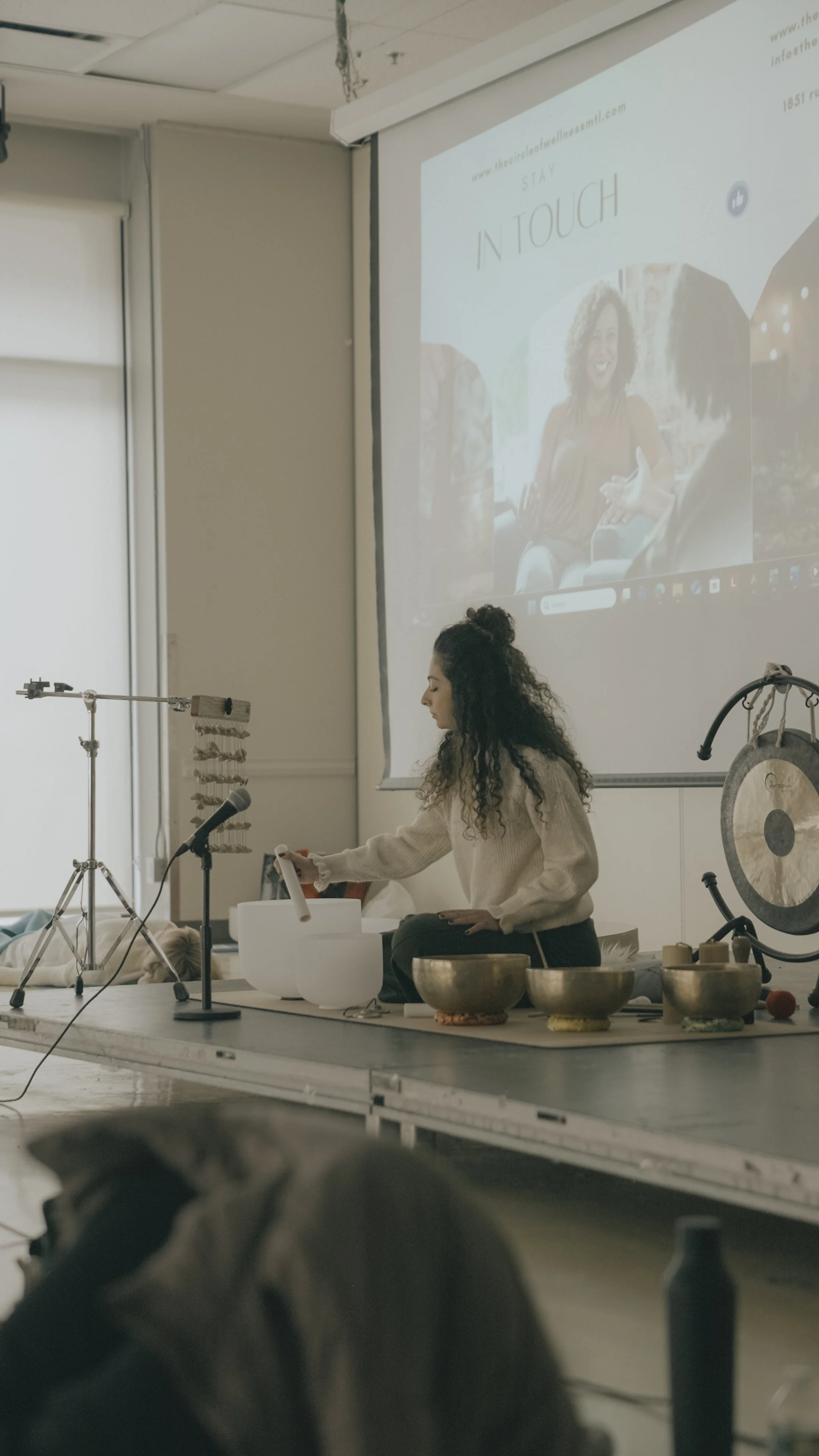 Woman sitting on a stage or platform performing a sound or meditation session with singing bowls and a mallet. A large gong is positioned nearby. A presentation slide with the words 'Stay In Touch' and a photo of a woman talking is projected on a screen behind her.