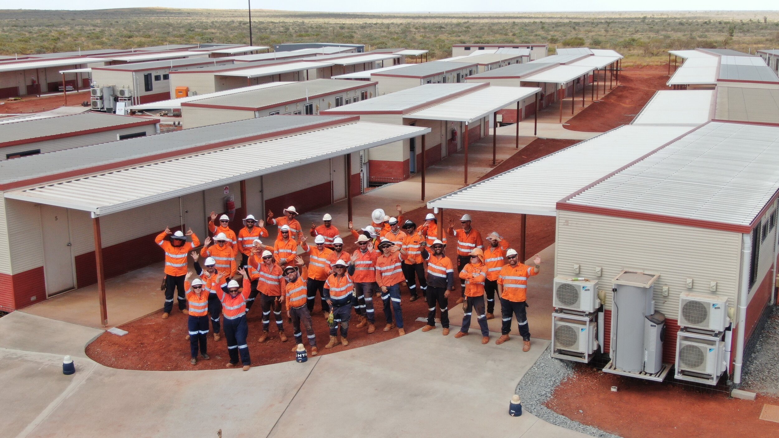 Group of construction workers in orange safety vests and white hard hats, standing outdoors in front of temporary modular buildings, waving and posing for the photo.