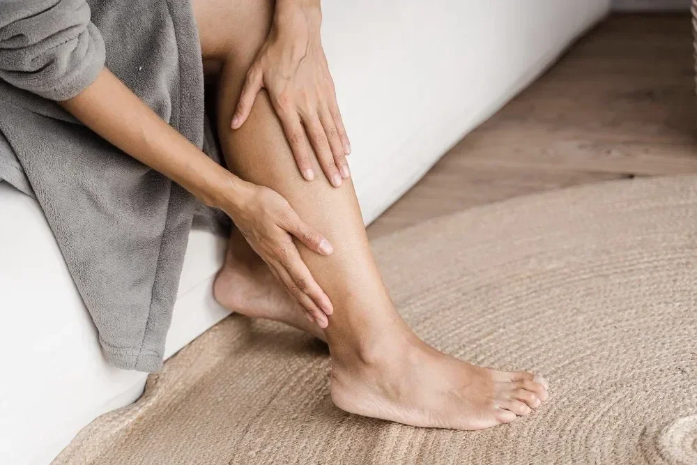 A person sitting on a white sofa is holding their lower leg and ankle, appearing to experience pain or discomfort.