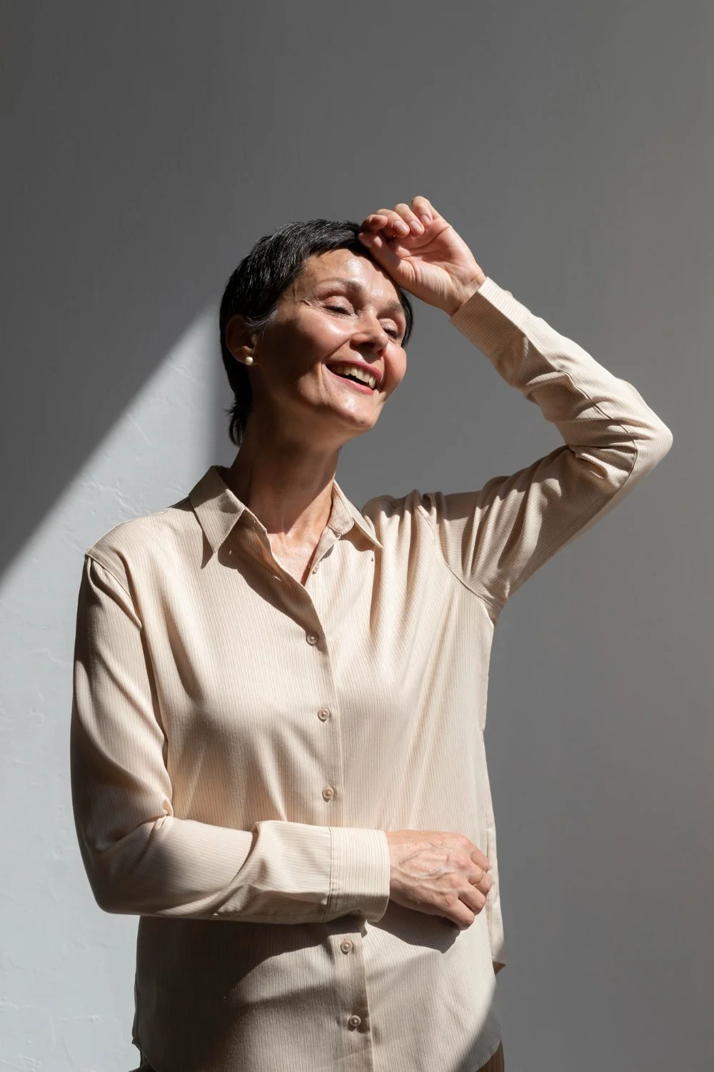 A happy older woman with short dark hair, wearing a beige button-up shirt, smiling with eyes closed, shielding her face from sunlight, standing against a plain background.