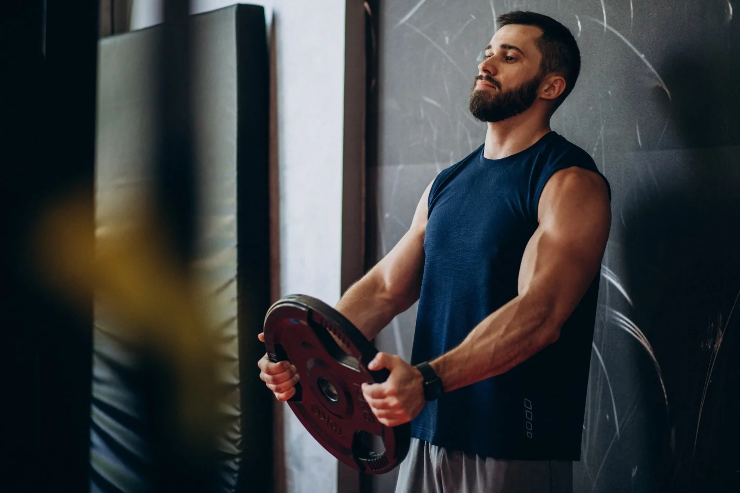 A man with a beard in a sleeveless navy blue shirt holding a weight plate in a gym, leaning against a dark chalkboard wall.