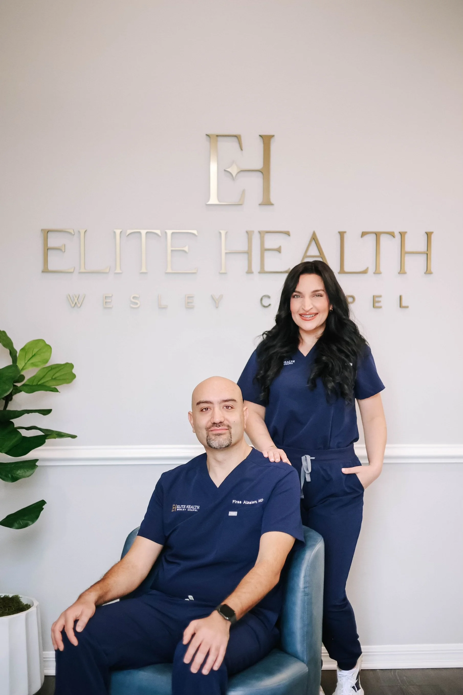 Two healthcare professionals in navy scrubs, a woman with long black hair standing and a man with a shaved head sitting in a chair, in front of a wall with 'Elite Health Wesley Chapel' signage.