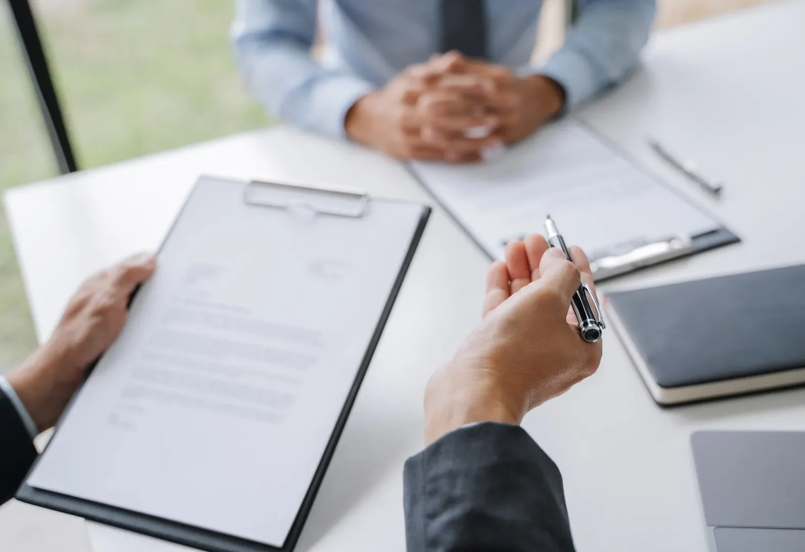Close-up of two people in a business meeting, with one person holding a clipboard and the other holding a pen over paper, at a white table with laptops and notebooks.