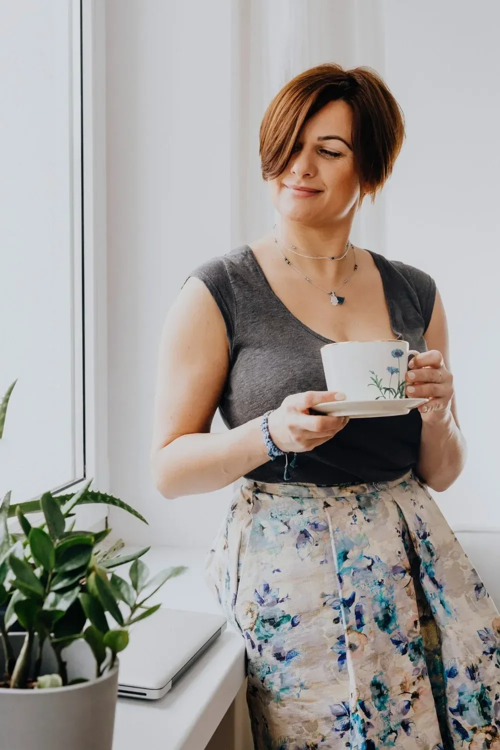 A woman with short brown hair, wearing a gray sleeveless top and floral beige pants, standing near a window and holding a white teacup with a floral design.