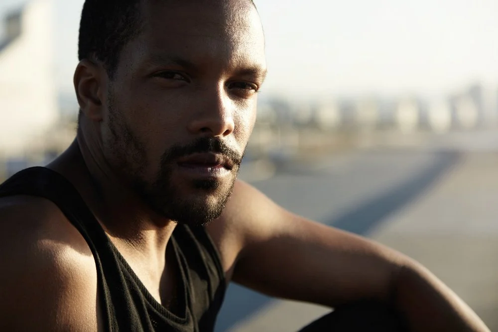 Close-up of a man with short black hair and a beard, wearing a black sleeveless shirt, looking at the camera with a serious expression, outdoors during daytime.
