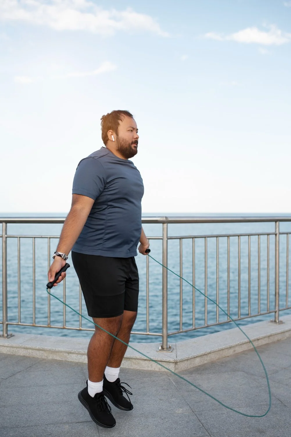 Man skipping rope near the water on a pier or promenade, with railing and open ocean in the background.