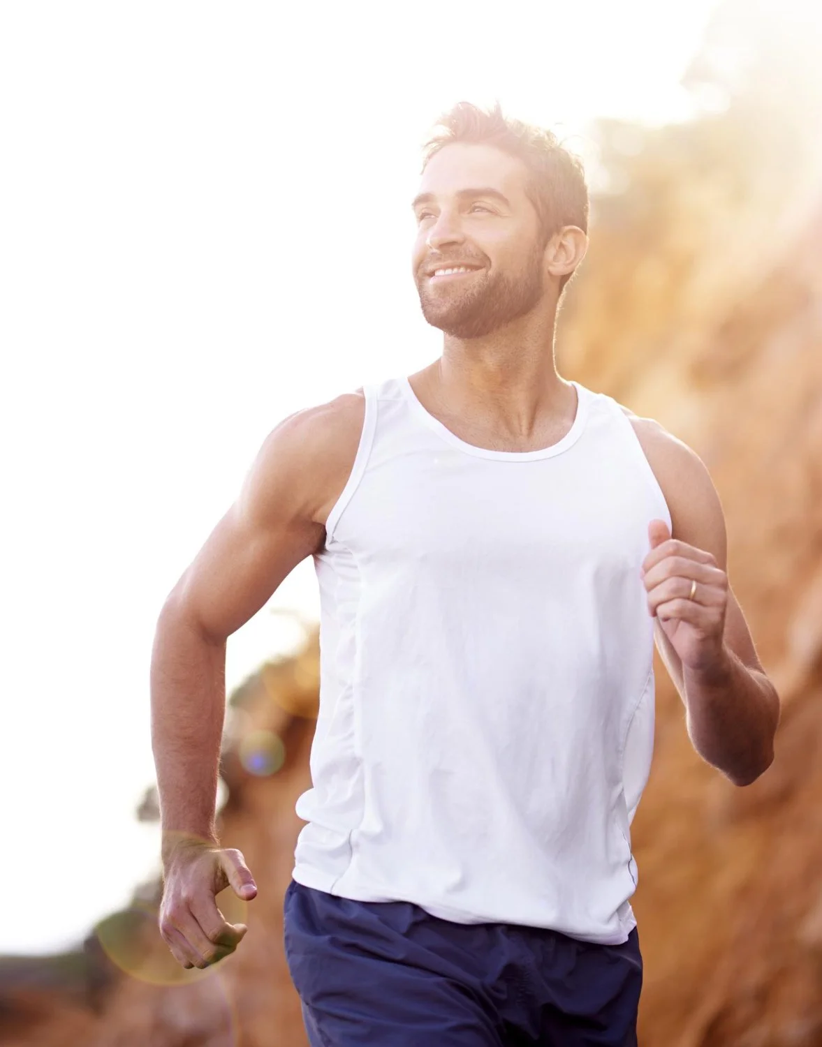 A man running outdoors in a park or trail during daylight, wearing a white tank top and navy shorts.