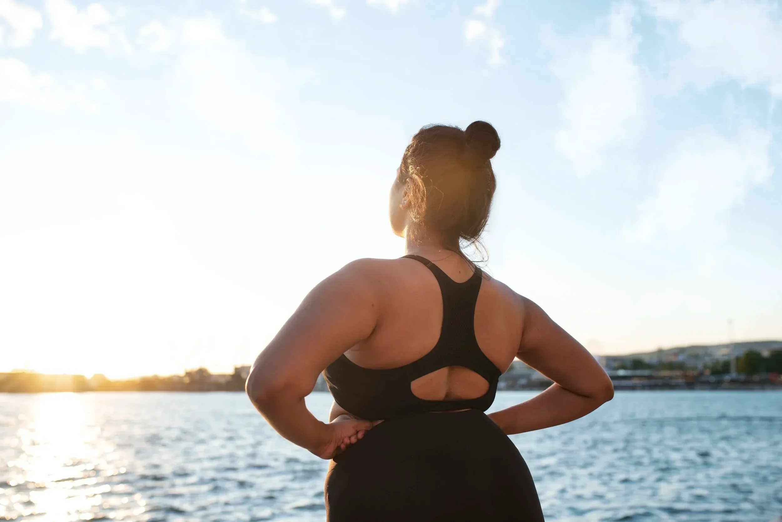 Woman in black athletic wear standing by the water at sunset, facing away from the camera with hands on hips.