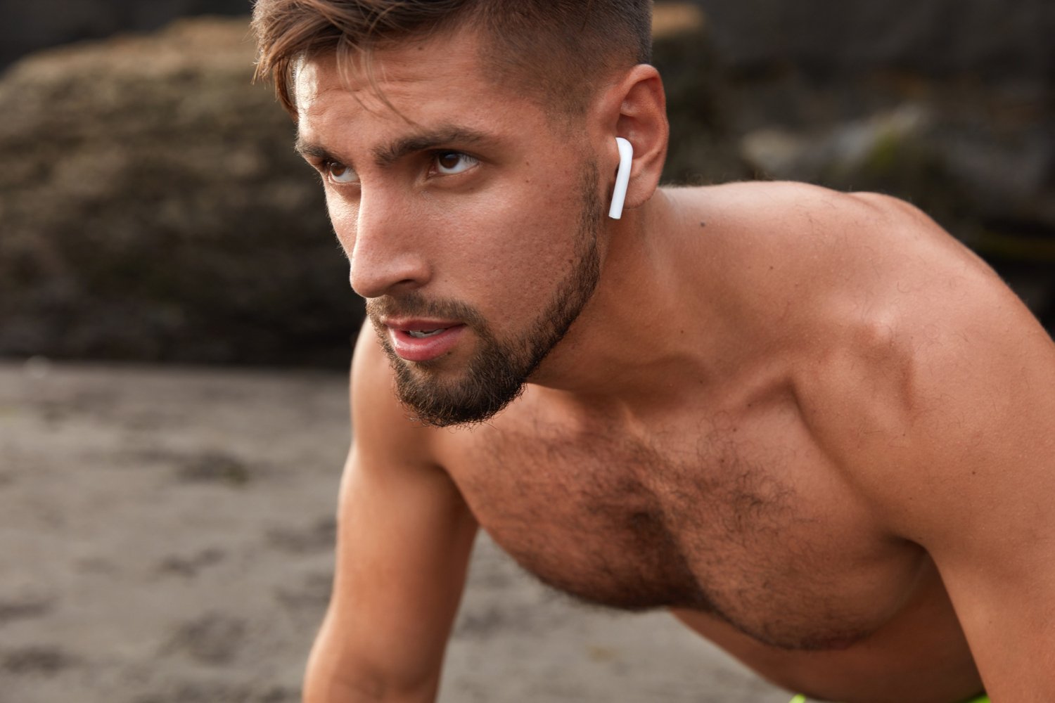 A shirtless man with a beard and short hair wearing wireless earbuds, outdoors near rocks.