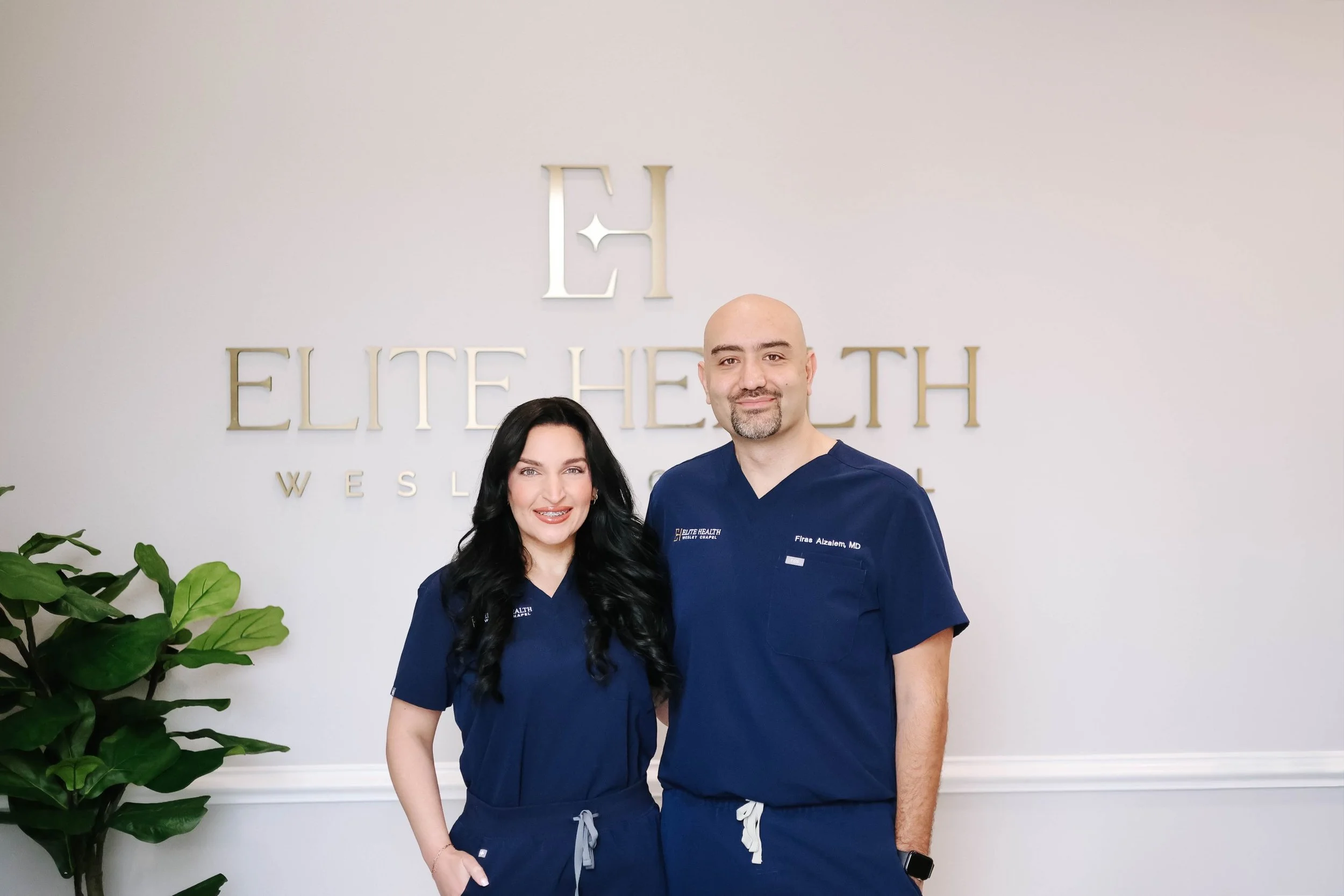 Dr. Firas Alzaiem and Nurse Wanda Khalil wearing blue medical scrubs standing in front of a wall with a sign that reads 'Elite Health' and a logo above it.